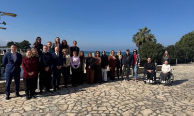 Na fotografiji je velika skupina od 22 ljudi koji poziraju na otvorenom, stojeći na kamenoj terasi. Iza njih se pruža prekrasan pogled na more, plavo nebo i zelenilo, uključujući i jednu visoku palmu koja daje mediteranski ugođaj.