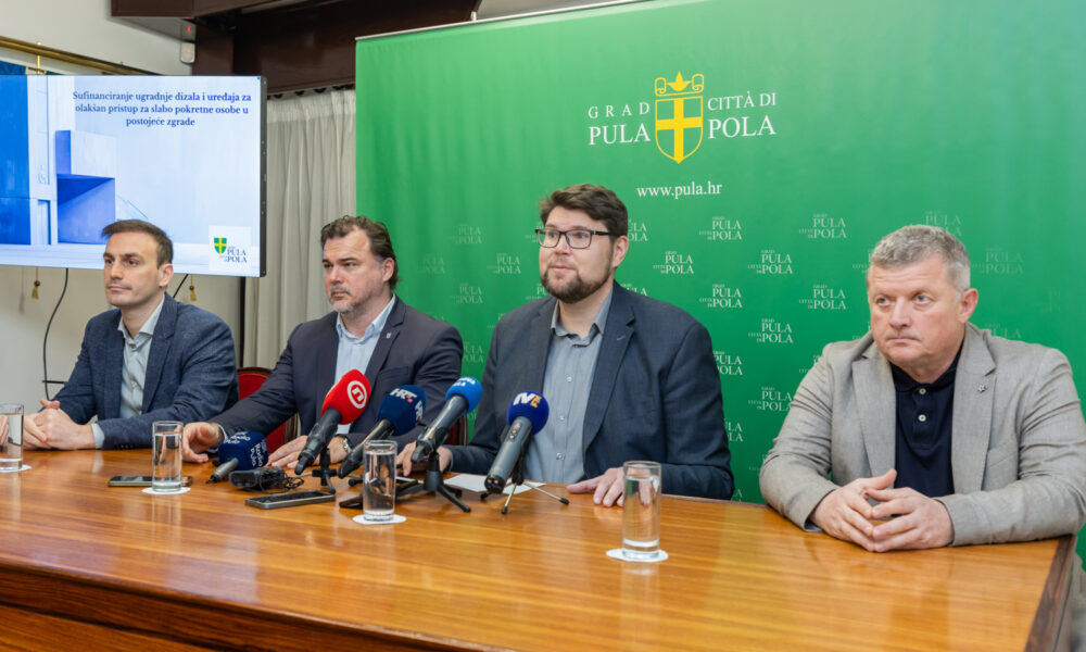 Na fotografiji se vidi službena konferencija za medije ili javno predstavljanje u organizaciji Grada Pule. Četvorica muškaraca sjede za drvenim stolom, ispred njih su mikrofoni i čaše vode, što upućuje na formalni događaj. Iza njih je velika zelena pozadina s grbom grada i natpisom “GRAD PULA – CITTÀ DI POLA” te službenom web‑adresom. Na lijevoj strani fotografije nalazi se projekcijsko platno s prikazanim naslovnim slajdom prezentacije: “Sufinanciranje ugradnje dizala i uređaja za olakšan pristup za slabo pokretne osobe u postojeće zgrade”. Tema je jasno vezana uz poboljšanje pristupačnosti i potporu osobama smanjene pokretljivosti.