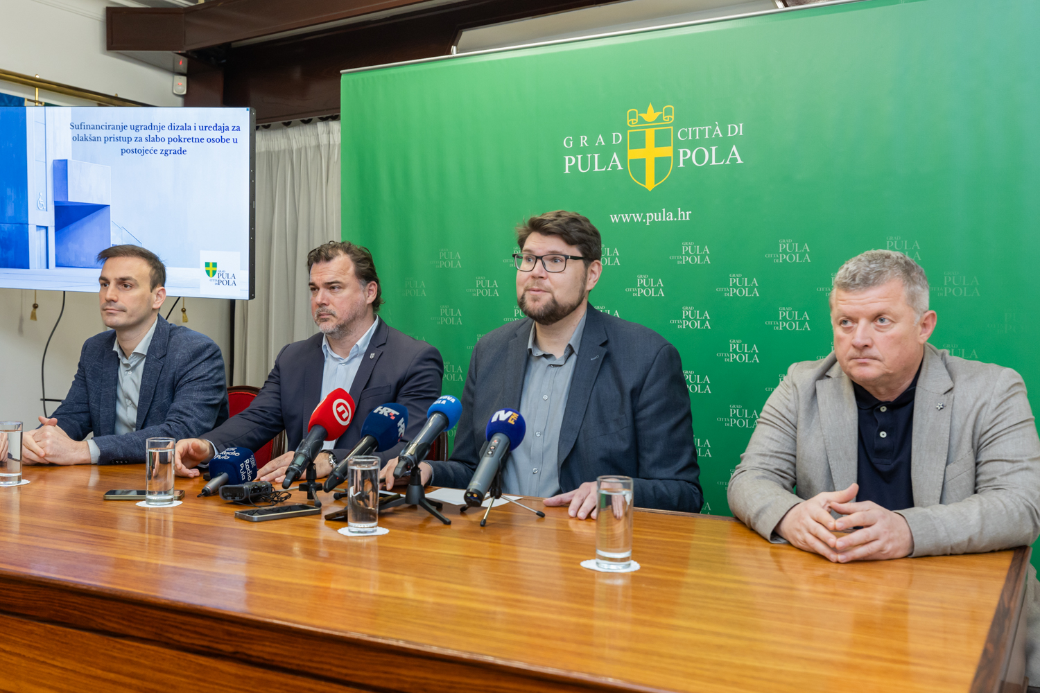 Na fotografiji se vidi službena konferencija za medije ili javno predstavljanje u organizaciji Grada Pule. Četvorica muškaraca sjede za drvenim stolom, ispred njih su mikrofoni i čaše vode, što upućuje na formalni događaj. Iza njih je velika zelena pozadina s grbom grada i natpisom “GRAD PULA – CITTÀ DI POLA” te službenom web‑adresom. Na lijevoj strani fotografije nalazi se projekcijsko platno s prikazanim naslovnim slajdom prezentacije: “Sufinanciranje ugradnje dizala i uređaja za olakšan pristup za slabo pokretne osobe u postojeće zgrade”. Tema je jasno vezana uz poboljšanje pristupačnosti i potporu osobama smanjene pokretljivosti.