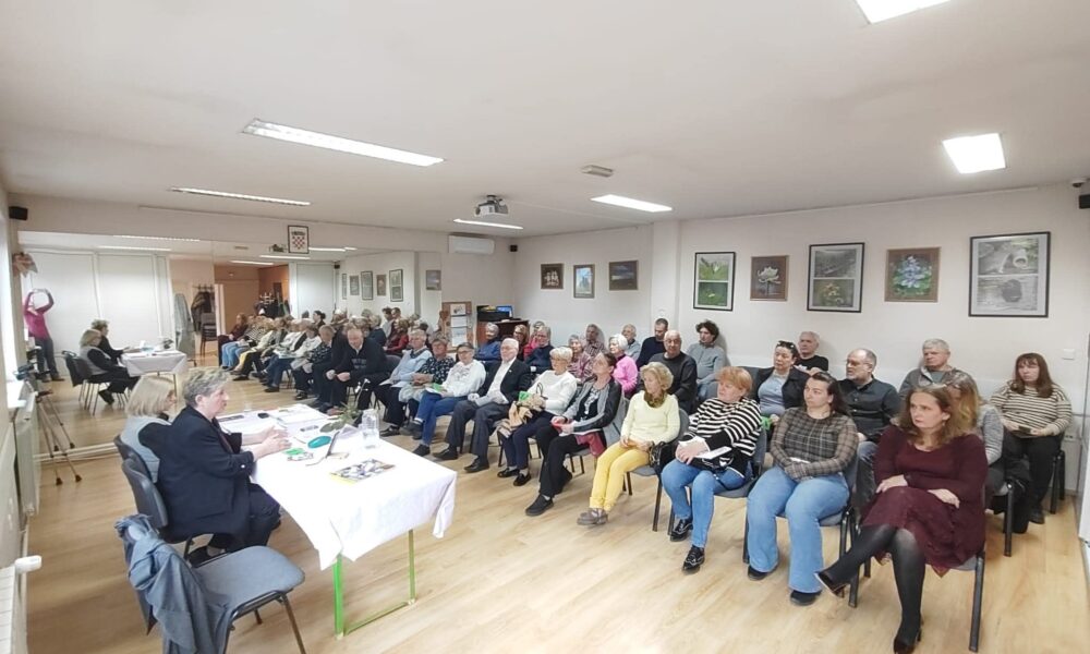 Fotografija prikazuje skup ili zajednički sastanak u svijetloj, urednoj prostoriji s drvenim podom. Oko četrdesetak ljudi sjedi u urednim redovima, svi okrenuti prema stolu na kojem sjede dvije osobe koje vode raspravu ili prezentaciju. Sudionici djeluju koncentrirano i uključeno, što stvara dojam ozbiljnog, ali konstruktivnog okupljanja. Na zidovima vise uokvirene fotografije ili umjetnička djela, a na stropu se nalazi projektor, što sugerira da se možda koriste i vizualni materijali. U prostoriji je postavljena i kamera na stativu, vjerojatno za snimanje ili prijenos događaja. Cijela scena odiše osjećajem zajedništva i aktivnog sudjelovanja u nekoj vrsti edukativnog ili društvenog programa.