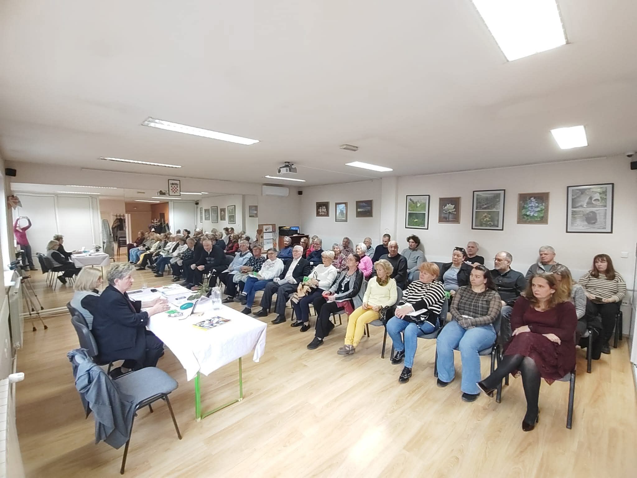 Fotografija prikazuje skup ili zajednički sastanak u svijetloj, urednoj prostoriji s drvenim podom. Oko četrdesetak ljudi sjedi u urednim redovima, svi okrenuti prema stolu na kojem sjede dvije osobe koje vode raspravu ili prezentaciju. Sudionici djeluju koncentrirano i uključeno, što stvara dojam ozbiljnog, ali konstruktivnog okupljanja. Na zidovima vise uokvirene fotografije ili umjetnička djela, a na stropu se nalazi projektor, što sugerira da se možda koriste i vizualni materijali. U prostoriji je postavljena i kamera na stativu, vjerojatno za snimanje ili prijenos događaja. Cijela scena odiše osjećajem zajedništva i aktivnog sudjelovanja u nekoj vrsti edukativnog ili društvenog programa.