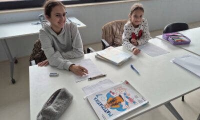 Na slici su dvoje djece koja sjede za bijelim stolom u učionici i rade na školskim zadacima. Oboje drže olovke i imaju ispred sebe prazne listove papira, kao da se pripremaju nešto napisati ili riješiti.