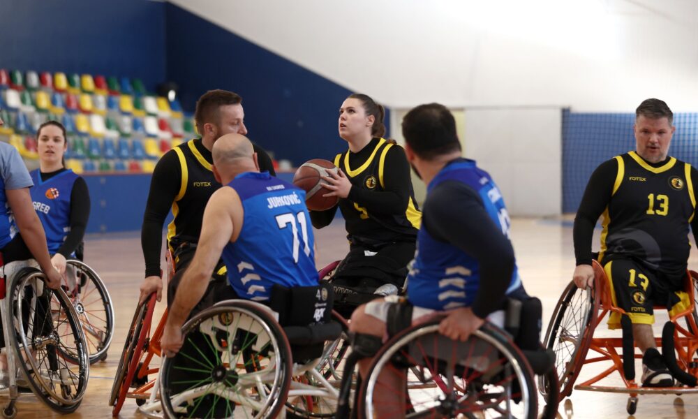 Fotografija prikazuje nekoliko igrača košarke u kolicima kako igraju košarku u kolicima. Igračica u kolicima pokušava pucati ali je okružena s više igrača, neki su iz njene, a neki iz protivničke momčadi.