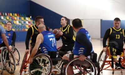 Fotografija prikazuje nekoliko igrača košarke u kolicima kako igraju košarku u kolicima. Igračica u kolicima pokušava pucati ali je okružena s više igrača, neki su iz njene, a neki iz protivničke momčadi.