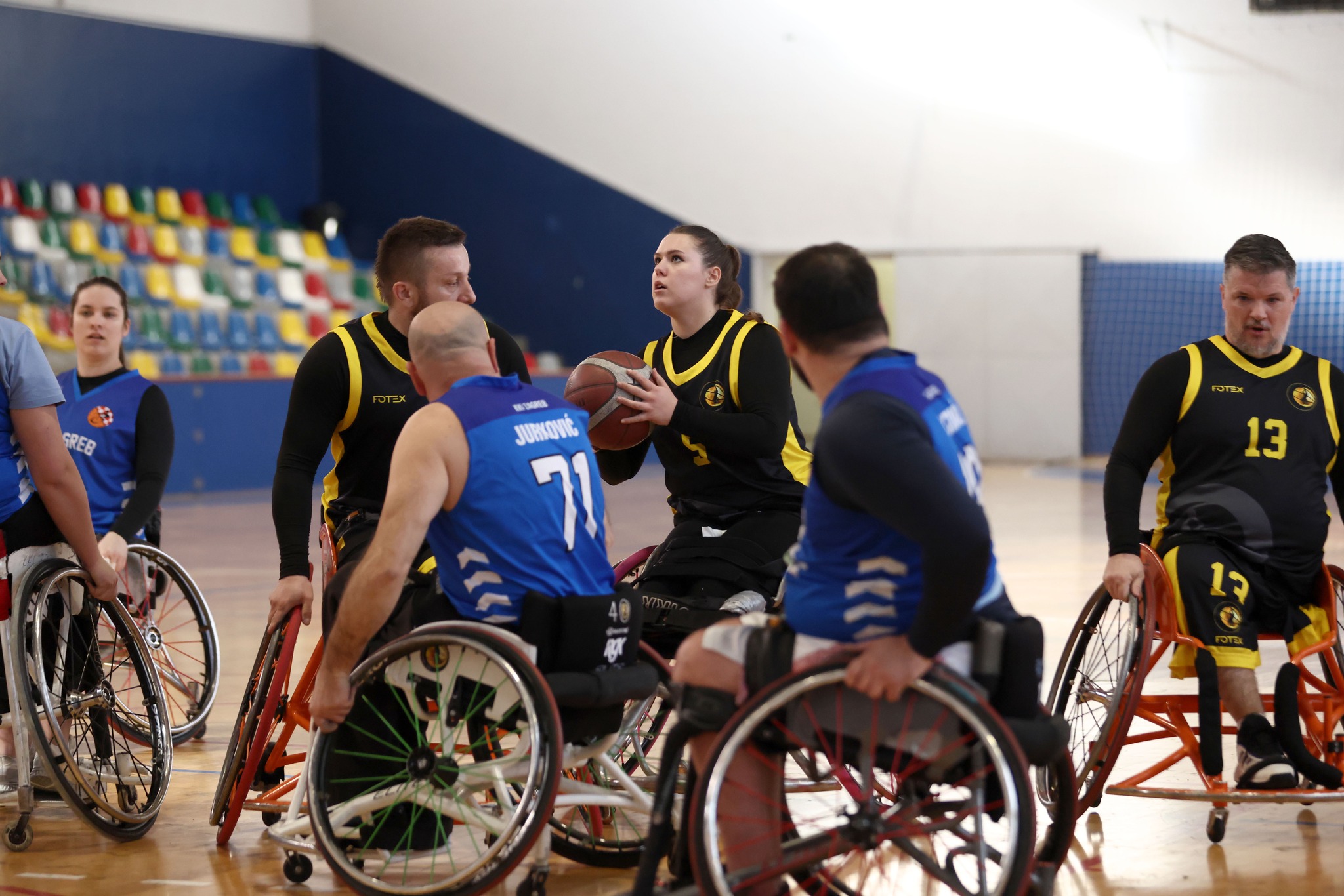 Fotografija prikazuje nekoliko igrača košarke u kolicima kako igraju košarku u kolicima. Igračica u kolicima pokušava pucati ali je okružena s više igrača, neki su iz njene, a neki iz protivničke momčadi.