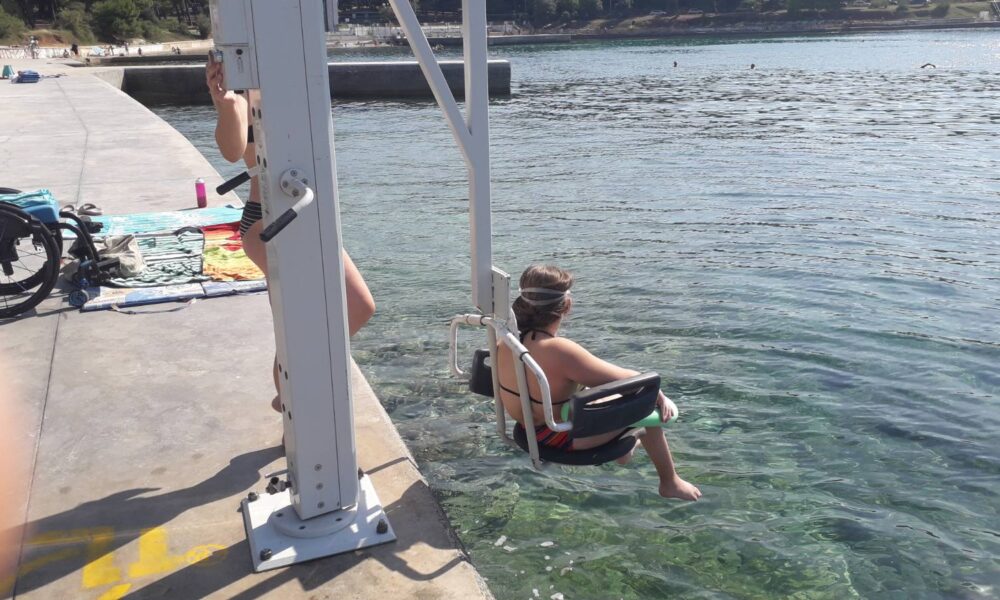 Fotografija prikazuje osobu koja uz pomoć mehaničke dizalice ulazi u more s betonskog mola. Ova vrsta dizalice služi za olakšavanje pristupa vodi osobama s ograničenom pokretljivošću. Na molu se vidi invalidska kolica, ručnik i nekoliko osobnih stvari, što upućuje na to da se osoba koja koristi dizalicu inače kreće u kolicima. U blizini stoji još jedna osoba, vjerojatno kako bi pomogla ili nadzirala postupak. More je mirno i bistro, a u pozadini se vide drugi kupači koji uživaju u plivanju. Cijela scena odiše inkluzivnošću i pokazuje kako prilagođena infrastruktura omogućuje svima da ravnopravno uživaju u ljetnim aktivnostima.