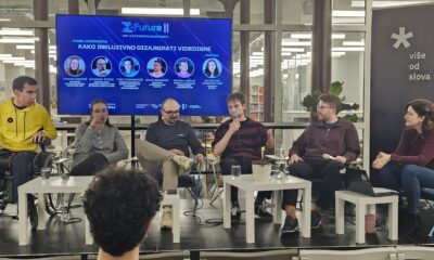 Na fotografiji se vidi panel‑rasprava održana u modernom, svijetlom prostoru, vjerojatno knjižnici ili konferencijskoj dvorani. Ispred velike digitalne pozadine sjedi šest sudionika u polukrugu. Svi su usmjereni prema publici i djeluju angažirano u razgovoru. Jedna osoba je u invalidskim kolicima. Tema panela je “Kako inkluzivno dizajnirati videoigre”, što naglašava fokus na pristupačnost i inkluziju u gaming industriji.