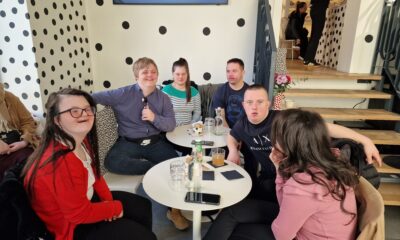 Na fotografiji se vidi živahan, moderan kafić uređen u upečatljivom stilu: bijele površine prekrivene velikim crnim točkama stvaraju razigranu, gotovo umjetničku atmosferu. U prvom planu sjedi sedam osoba oko dva mala okrugla stola. Na stolovima su čaše, boce i pića, a ljudi djeluju opušteno — neki razgovaraju, neki gledaju prema kameri. Pozadina otkriva stepenište koje vodi na gornju etažu, gdje se vide još dvije osobe. Cijeli prostor izgleda kao moderno, društveno mjesto, možda popularan tematski kafić ili restoran. Dojam je vrlo prijateljski i ležeran, kao da je riječ o ugodnom druženju u zanimljivo uređenom prostoru.