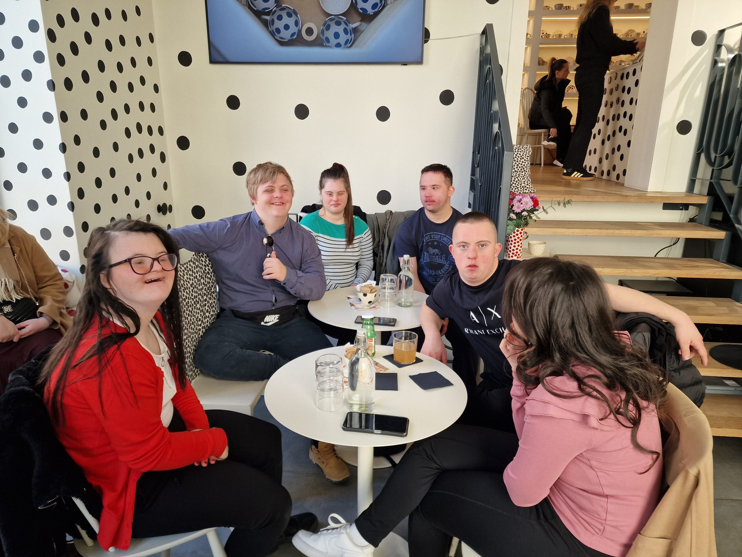 Na fotografiji se vidi živahan, moderan kafić uređen u upečatljivom stilu: bijele površine prekrivene velikim crnim točkama stvaraju razigranu, gotovo umjetničku atmosferu. U prvom planu sjedi sedam osoba oko dva mala okrugla stola. Na stolovima su čaše, boce i pića, a ljudi djeluju opušteno — neki razgovaraju, neki gledaju prema kameri. Pozadina otkriva stepenište koje vodi na gornju etažu, gdje se vide još dvije osobe. Cijeli prostor izgleda kao moderno, društveno mjesto, možda popularan tematski kafić ili restoran. Dojam je vrlo prijateljski i ležeran, kao da je riječ o ugodnom druženju u zanimljivo uređenom prostoru.