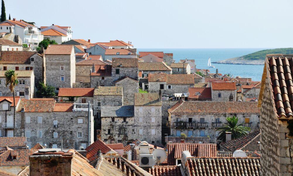 Na fotografiji je prikazan slikovit pogled na stari dio grada Hvara. Dominiraju kamene kuće zbijene jedna uz drugu, s karakterističnim crvenim krovovima od crijepa koji stvaraju topao, mediteranski ugođaj. Građevine su različitih visina i stilova, ali sve dijele tradicionalnu dalmatinsku arhitekturu – kamene fasade, male prozore i ponegdje drvene škure. Između kuća proviruju palme i zelenilo, što dodatno naglašava primorski karakter mjesta. U pozadini se vidi more mirne plave boje, a na horizontu i mali jedrilica koja plovi uz obalu. S desne strane nazire se i dio otoka ili kopna, što daje osjećaj širine i otvorenosti prostora. Cijela scena odiše autentičnošću, poviješću i opuštenom atmosferom tipičnom za dalmatinske gradove.