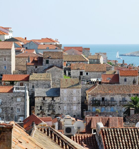 Na fotografiji je prikazan slikovit pogled na stari dio grada Hvara. Dominiraju kamene kuće zbijene jedna uz drugu, s karakterističnim crvenim krovovima od crijepa koji stvaraju topao, mediteranski ugođaj. Građevine su različitih visina i stilova, ali sve dijele tradicionalnu dalmatinsku arhitekturu – kamene fasade, male prozore i ponegdje drvene škure. Između kuća proviruju palme i zelenilo, što dodatno naglašava primorski karakter mjesta. U pozadini se vidi more mirne plave boje, a na horizontu i mali jedrilica koja plovi uz obalu. S desne strane nazire se i dio otoka ili kopna, što daje osjećaj širine i otvorenosti prostora. Cijela scena odiše autentičnošću, poviješću i opuštenom atmosferom tipičnom za dalmatinske gradove.