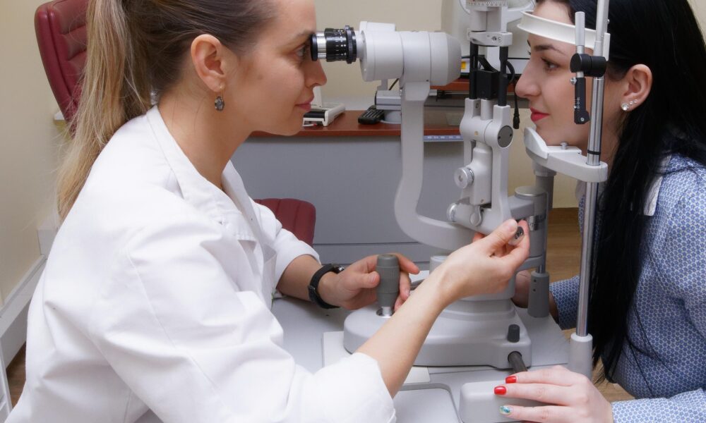 Na fotografiji se vidi pregled vida u oftalmološkoj ordinaciji. Pacijentica sjedi ispred rasvjetne biomikroskopske lampe (slit lampe), s bradom i čelom naslonjenima na držače uređaja. Oftalmolog ili optometrist pažljivo promatra njezino oko kroz okular i podešava instrument kako bi dobio bolji uvid u stanje oka. U pozadini se nalazi dodatna oftalmološka oprema, uključujući foropter, uređaj koji se koristi za određivanje dioptrije. Cijela scena djeluje profesionalno i smireno, tipična za rutinski pregled vida.