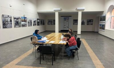 Na fotografiji se vidi četvero ljudi koji sjede oko pravokutnog drvenog stola u prostranoj prostoriji s bijelim zidovima i pločicama na podu. Svi su zaokupljeni radom: pred njima su bilježnice, papiri, olovke i jedna torba. S lijeve strane sjedi jedna osoba sama, dok su s desne strane troje smješteni bliže jedno drugome, kao mala radna skupina. Zidovi prostorije prekriveni su brojnim uokvirenim fotografijama koje prikazuju zgrade, arhitekturu i prizore koji djeluju povijesno ili dokumentarno — moguće i prizore razaranja. U stražnjem dijelu prostorije nalazi se i veliki pano s natpisom "REFERENDUM", što sugerira da se radi o muzeju, kulturnom centru ili izložbenom prostoru s političkom ili povijesnom tematikom. Atmosfera djeluje mirno i fokusirano, kao da se u prostoru uči, istražuje ili priprema neki projekt.