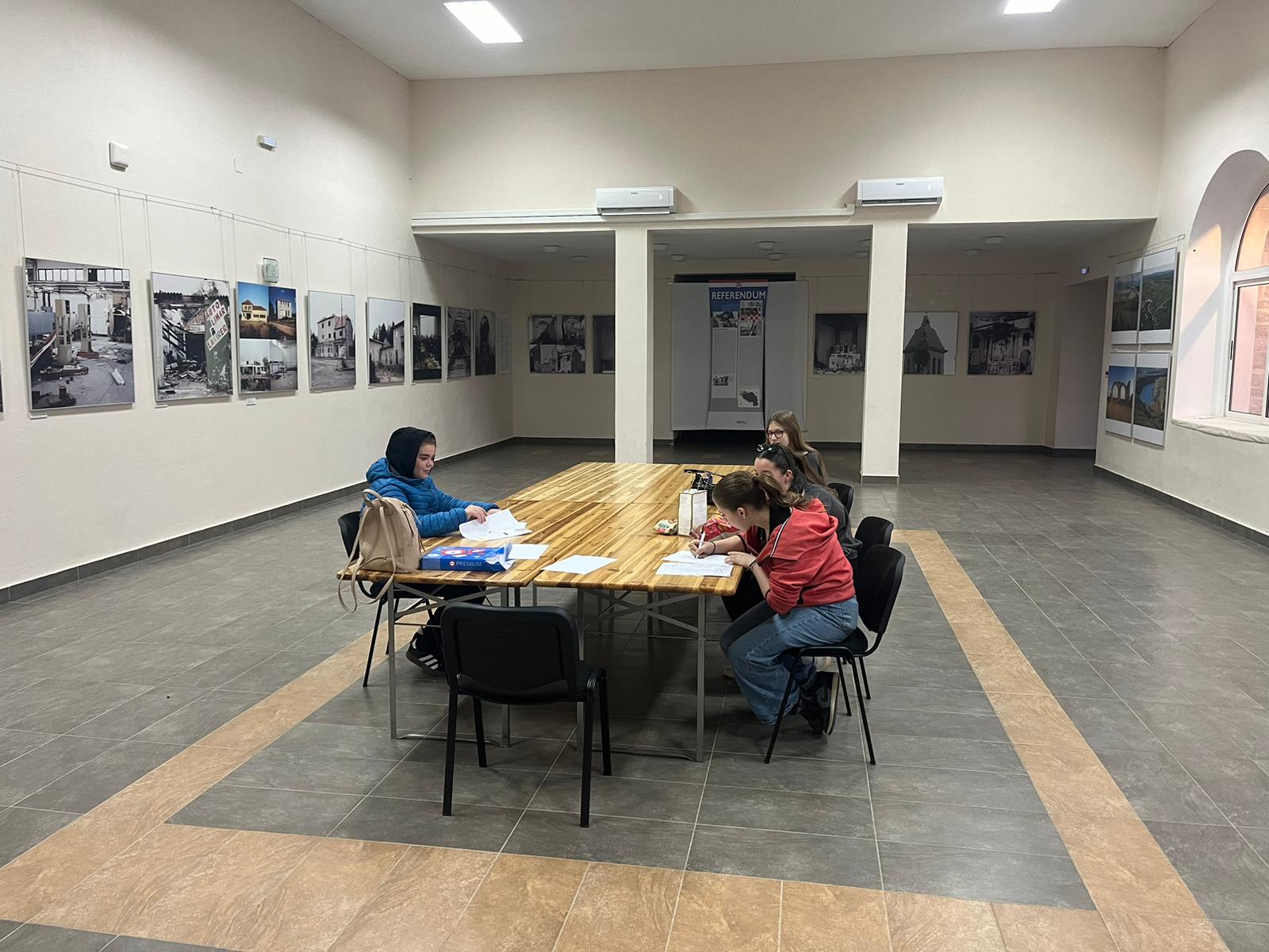 Na fotografiji se vidi četvero ljudi koji sjede oko pravokutnog drvenog stola u prostranoj prostoriji s bijelim zidovima i pločicama na podu. Svi su zaokupljeni radom: pred njima su bilježnice, papiri, olovke i jedna torba. S lijeve strane sjedi jedna osoba sama, dok su s desne strane troje smješteni bliže jedno drugome, kao mala radna skupina. Zidovi prostorije prekriveni su brojnim uokvirenim fotografijama koje prikazuju zgrade, arhitekturu i prizore koji djeluju povijesno ili dokumentarno — moguće i prizore razaranja. U stražnjem dijelu prostorije nalazi se i veliki pano s natpisom "REFERENDUM", što sugerira da se radi o muzeju, kulturnom centru ili izložbenom prostoru s političkom ili povijesnom tematikom. Atmosfera djeluje mirno i fokusirano, kao da se u prostoru uči, istražuje ili priprema neki projekt.
