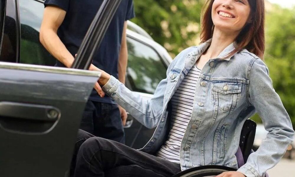 Na fotografiji se vidi žena koja sjedi u invalidskim kolicima i s osmijehom izlazi iz automobila. Odjevena je ležerno, u traper jaknu i prugastu majicu, a njezin izraz lica odaje zadovoljstvo i dobro raspoloženje. Jednom rukom drži vrata automobila, dok drugom pridržava kolica. Uz nju stoji druga osoba, vjerojatno pomagač ili pratnja, koja joj asistira pri izlasku iz vozila. Automobil je parkiran na otvorenom, a u pozadini se vidi zelenilo, što sugerira da se scena odvija u prirodnom ili urbanom okruženju s puno drveća. Cijela scena djeluje pozitivno i podržavajuće, naglašavajući samostalnost, ali i važnost pomoći i pristupačnosti u svakodnevnim situacijama.
