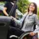 Na fotografiji se vidi žena koja sjedi u invalidskim kolicima i s osmijehom izlazi iz automobila. Odjevena je ležerno, u traper jaknu i prugastu majicu, a njezin izraz lica odaje zadovoljstvo i dobro raspoloženje. Jednom rukom drži vrata automobila, dok drugom pridržava kolica. Uz nju stoji druga osoba, vjerojatno pomagač ili pratnja, koja joj asistira pri izlasku iz vozila. Automobil je parkiran na otvorenom, a u pozadini se vidi zelenilo, što sugerira da se scena odvija u prirodnom ili urbanom okruženju s puno drveća. Cijela scena djeluje pozitivno i podržavajuće, naglašavajući samostalnost, ali i važnost pomoći i pristupačnosti u svakodnevnim situacijama.