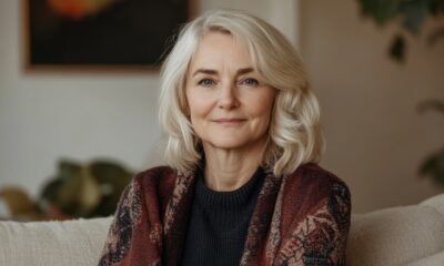 Na fotografiji je starija osoba sijede, duže kose, koja sjedi u udobnom, svijetlom dnevnom boravku. Odjevena je u tamniji pulover preko kojeg je prebačen šareni, topli šal u zemljanim tonovima. Sjedi na svijetloj sofi, opušteno i mirno, s blagim izrazom lica.