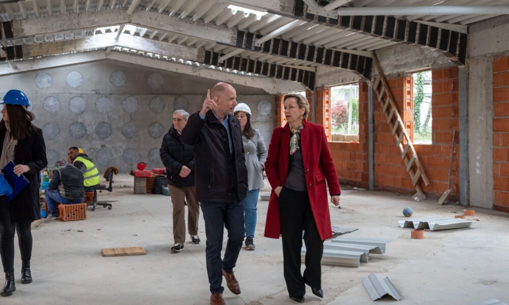 Grupa ljudi obilazi gradilište; muškarac objašnjava dok žena u crvenom kaputu sluša, radnici i građevinski materijal u pozadini.