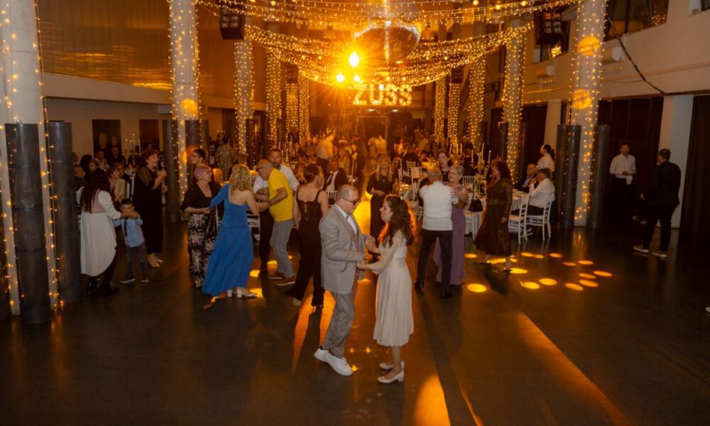 Na fotografiji je prikazana svečana večer u velikoj, elegantno uređenoj dvorani. Prostor je ukrašen toplim, zlatnim svjetlima koja vise sa stropa i stvaraju ugodnu i svečanu atmosferu. U središtu dvorane nalazi se plesni podij na kojem više parova pleše, dok se oko njih nalaze stolovi za kojima sjede i razgovaraju uzvanici. U prvom planu vidi se par koji pleše – muškarac u svijetlom odijelu i žena u svijetloj haljini, oboje opušteni i nasmijani. Oko njih su drugi gosti različitih generacija, od djece do starijih osoba, što daje dojam zajedništva i druženja. Ljudi su elegantno odjeveni, a atmosfera djeluje veselo i opušteno. U pozadini se nazire pozornica ili prostor za glazbu, dodatno naglašen svjetlosnim efektima. Cijela scena odiše toplinom, slavljem i dobrim raspoloženjem, tipičnim za svečane i humanitarne događaje.