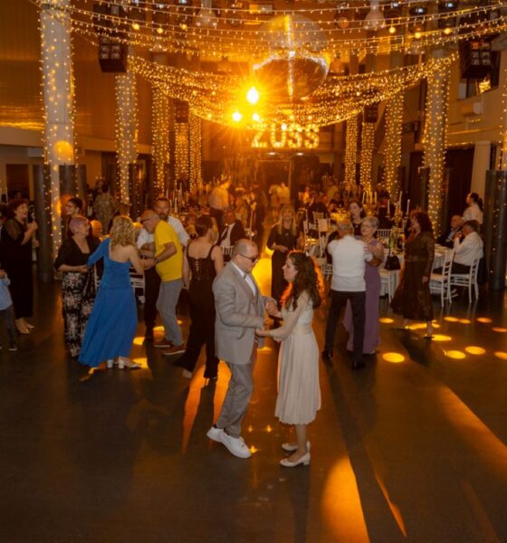 Na fotografiji je prikazana svečana večer u velikoj, elegantno uređenoj dvorani. Prostor je ukrašen toplim, zlatnim svjetlima koja vise sa stropa i stvaraju ugodnu i svečanu atmosferu. U središtu dvorane nalazi se plesni podij na kojem više parova pleše, dok se oko njih nalaze stolovi za kojima sjede i razgovaraju uzvanici. U prvom planu vidi se par koji pleše – muškarac u svijetlom odijelu i žena u svijetloj haljini, oboje opušteni i nasmijani. Oko njih su drugi gosti različitih generacija, od djece do starijih osoba, što daje dojam zajedništva i druženja. Ljudi su elegantno odjeveni, a atmosfera djeluje veselo i opušteno. U pozadini se nazire pozornica ili prostor za glazbu, dodatno naglašen svjetlosnim efektima. Cijela scena odiše toplinom, slavljem i dobrim raspoloženjem, tipičnim za svečane i humanitarne događaje.