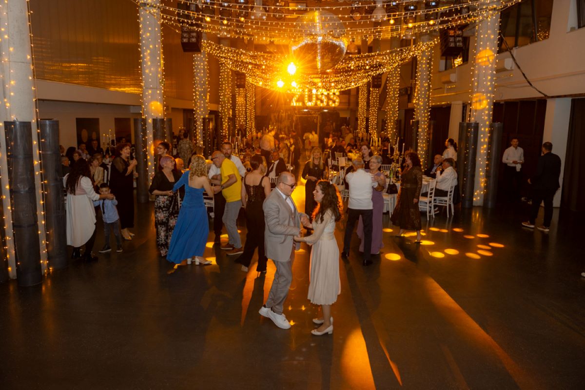 Na fotografiji je prikazana svečana večer u velikoj, elegantno uređenoj dvorani. Prostor je ukrašen toplim, zlatnim svjetlima koja vise sa stropa i stvaraju ugodnu i svečanu atmosferu. U središtu dvorane nalazi se plesni podij na kojem više parova pleše, dok se oko njih nalaze stolovi za kojima sjede i razgovaraju uzvanici. U prvom planu vidi se par koji pleše – muškarac u svijetlom odijelu i žena u svijetloj haljini, oboje opušteni i nasmijani. Oko njih su drugi gosti različitih generacija, od djece do starijih osoba, što daje dojam zajedništva i druženja. Ljudi su elegantno odjeveni, a atmosfera djeluje veselo i opušteno. U pozadini se nazire pozornica ili prostor za glazbu, dodatno naglašen svjetlosnim efektima. Cijela scena odiše toplinom, slavljem i dobrim raspoloženjem, tipičnim za svečane i humanitarne događaje.