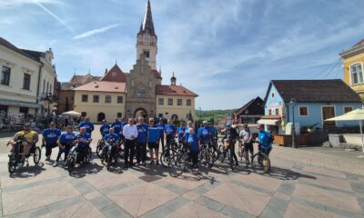 Na fotografiji se nalazi skupina ljudi okupljena na gradskom trgu, ispred povijesne zgrade s tornjem i satom. Riječ je o grupi sudionika biciklijade – neki stoje uz klasične bicikle, dok su drugi na prilagođenim biciklima za osobe s invaliditetom, uključujući ručne bicikle. Većina sudionika nosi plave majice, što sugerira da pripadaju istoj organizaciji ili događaju. U sredini skupine nalazi se nekoliko osoba koje poziraju zajedno, dok svi sudionici djeluju raspoloženo i spremno za zajedničku aktivnost. Okruženje je sunčano, s vedrim nebom i ugodnom atmosferom. U pozadini se vide šarene kuće, kafići i uređeni trg, što upućuje na turistički i kulturno značajan prostor. Fotografija odaje dojam zajedništva, inkluzije i pozitivne energije prije početka ili tijekom biciklijade.