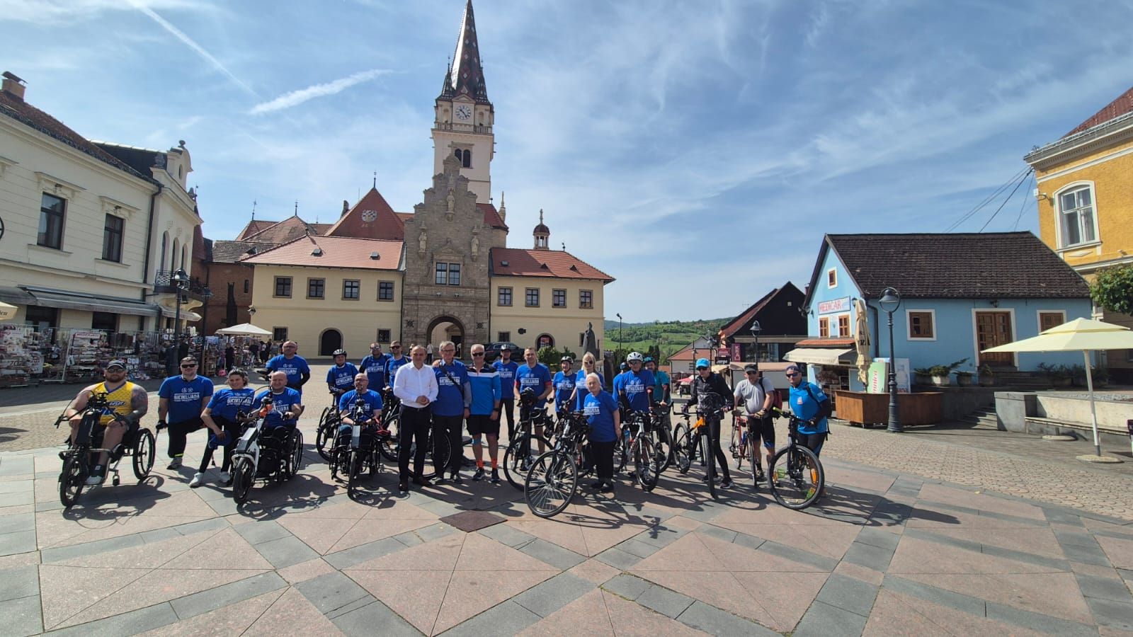 Na fotografiji se nalazi skupina ljudi okupljena na gradskom trgu, ispred povijesne zgrade s tornjem i satom. Riječ je o grupi sudionika biciklijade – neki stoje uz klasične bicikle, dok su drugi na prilagođenim biciklima za osobe s invaliditetom, uključujući ručne bicikle. Većina sudionika nosi plave majice, što sugerira da pripadaju istoj organizaciji ili događaju. U sredini skupine nalazi se nekoliko osoba koje poziraju zajedno, dok svi sudionici djeluju raspoloženo i spremno za zajedničku aktivnost. Okruženje je sunčano, s vedrim nebom i ugodnom atmosferom. U pozadini se vide šarene kuće, kafići i uređeni trg, što upućuje na turistički i kulturno značajan prostor. Fotografija odaje dojam zajedništva, inkluzije i pozitivne energije prije početka ili tijekom biciklijade.