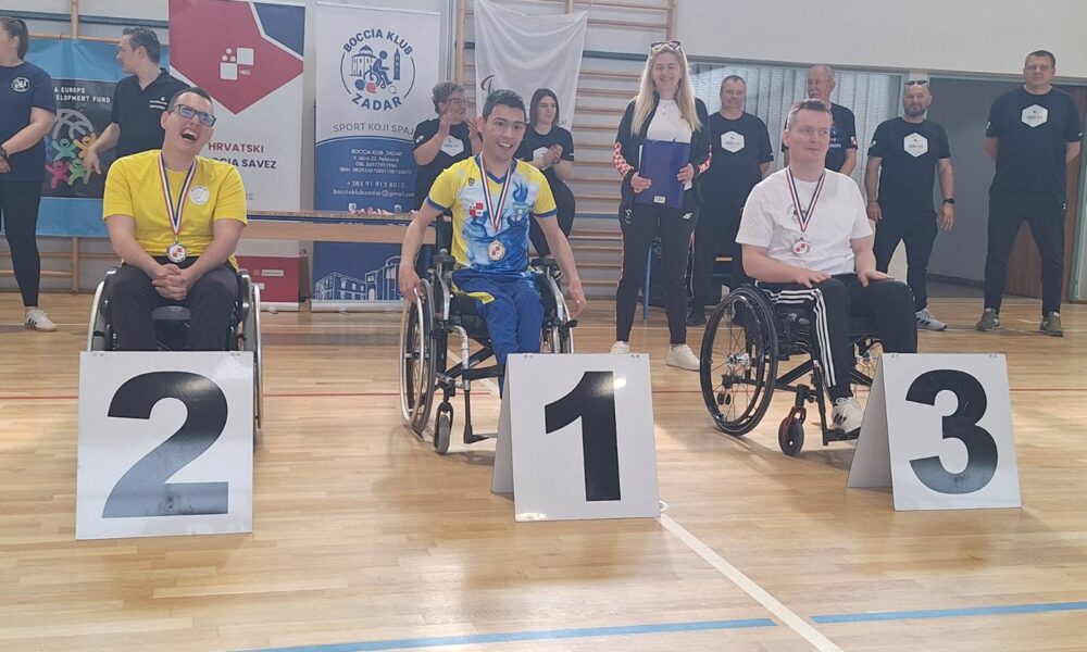 Fotografija prikazuje tri sportaša u kolicima s medaljama oko vrata, dok je ispred njih redni broj mjesta kojeg su osvojili.