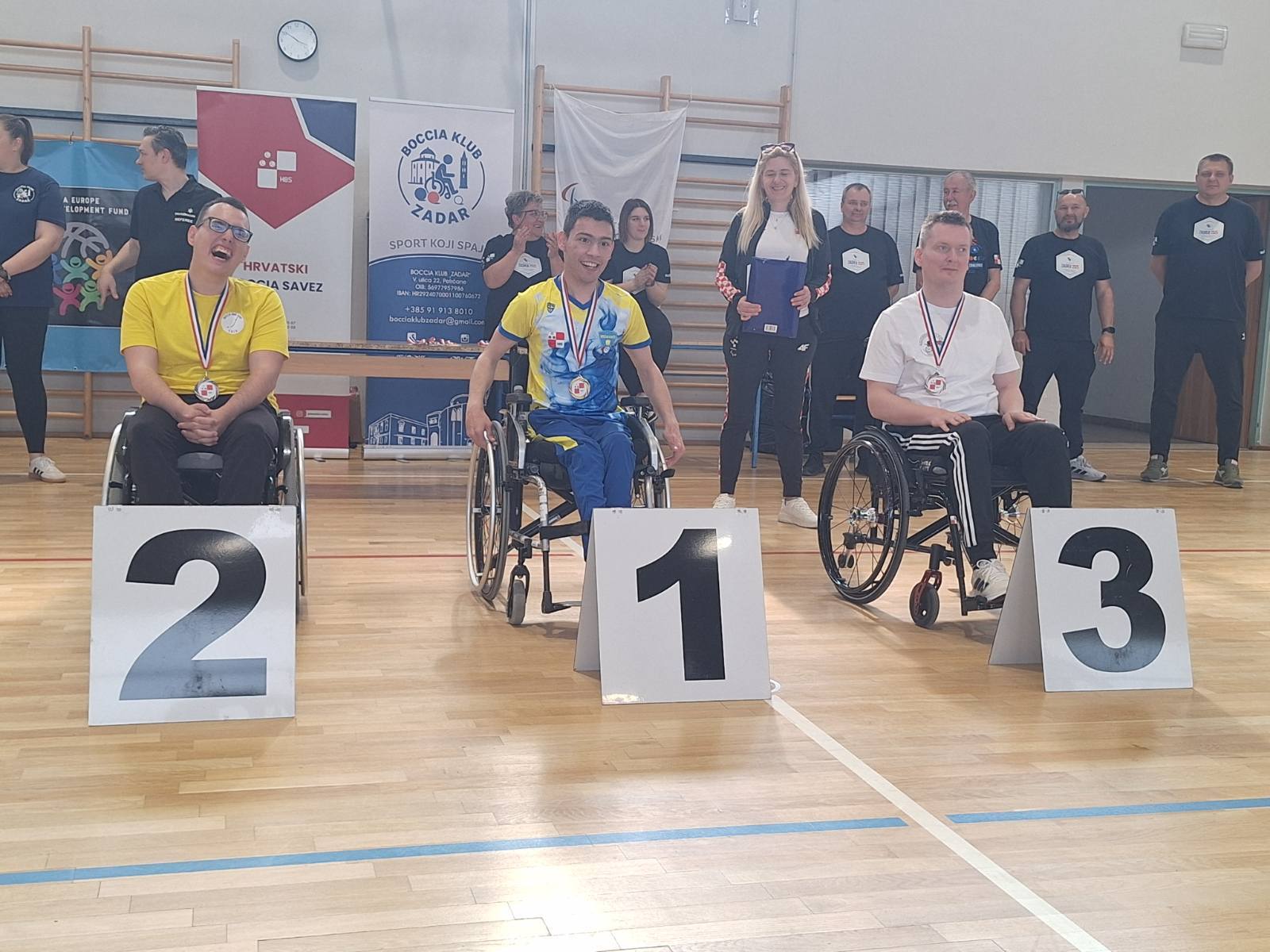 Fotografija prikazuje tri sportaša u kolicima s medaljama oko vrata, dok je ispred njih redni broj mjesta kojeg su osvojili.