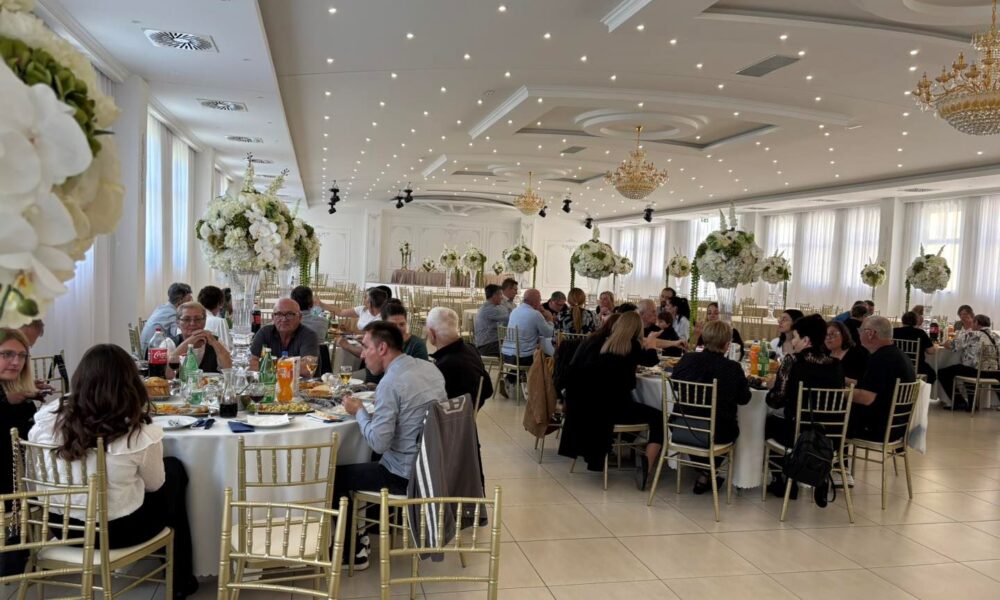 Na slici je prikazana svečana proslava u elegantnoj dvorani. Prostor je svijetao i prostran, s bijelim zidovima, visokim stropom ukrašenim rasvjetom i velikim lusterima koji dodatno naglašavaju svečani ugođaj. U dvorani se nalazi više okruglih stolova za kojima sjede uzvanici, većinom odrasli i stariji ljudi, odjeveni u polusvečanu i svečanu odjeću. Na stolovima su poslužena jela i pića, uključujući bezalkoholna pića, vino i vodu, što upućuje na organizirani ručak ili domjenak. Stolovi su dekorirani visokim cvjetnim aranžmanima u bijelim i zelenim tonovima, što prostoru daje profinjen i svečan izgled. Stolice su zlatne, a cijeli ambijent djeluje uredno i luksuzno. Ljudi za stolovima razgovaraju, jedu i druže se, što stvara dojam ugodne i opuštene atmosfere tijekom proslave ili obilježavanja nekog važnog događaja.