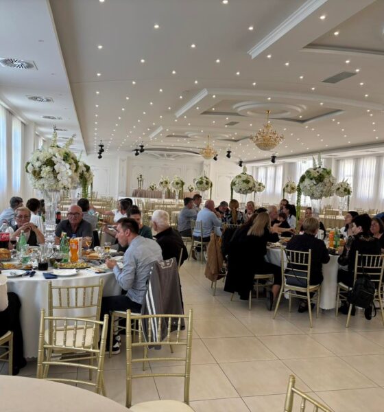 Na slici je prikazana svečana proslava u elegantnoj dvorani. Prostor je svijetao i prostran, s bijelim zidovima, visokim stropom ukrašenim rasvjetom i velikim lusterima koji dodatno naglašavaju svečani ugođaj. U dvorani se nalazi više okruglih stolova za kojima sjede uzvanici, većinom odrasli i stariji ljudi, odjeveni u polusvečanu i svečanu odjeću. Na stolovima su poslužena jela i pića, uključujući bezalkoholna pića, vino i vodu, što upućuje na organizirani ručak ili domjenak. Stolovi su dekorirani visokim cvjetnim aranžmanima u bijelim i zelenim tonovima, što prostoru daje profinjen i svečan izgled. Stolice su zlatne, a cijeli ambijent djeluje uredno i luksuzno. Ljudi za stolovima razgovaraju, jedu i druže se, što stvara dojam ugodne i opuštene atmosfere tijekom proslave ili obilježavanja nekog važnog događaja.