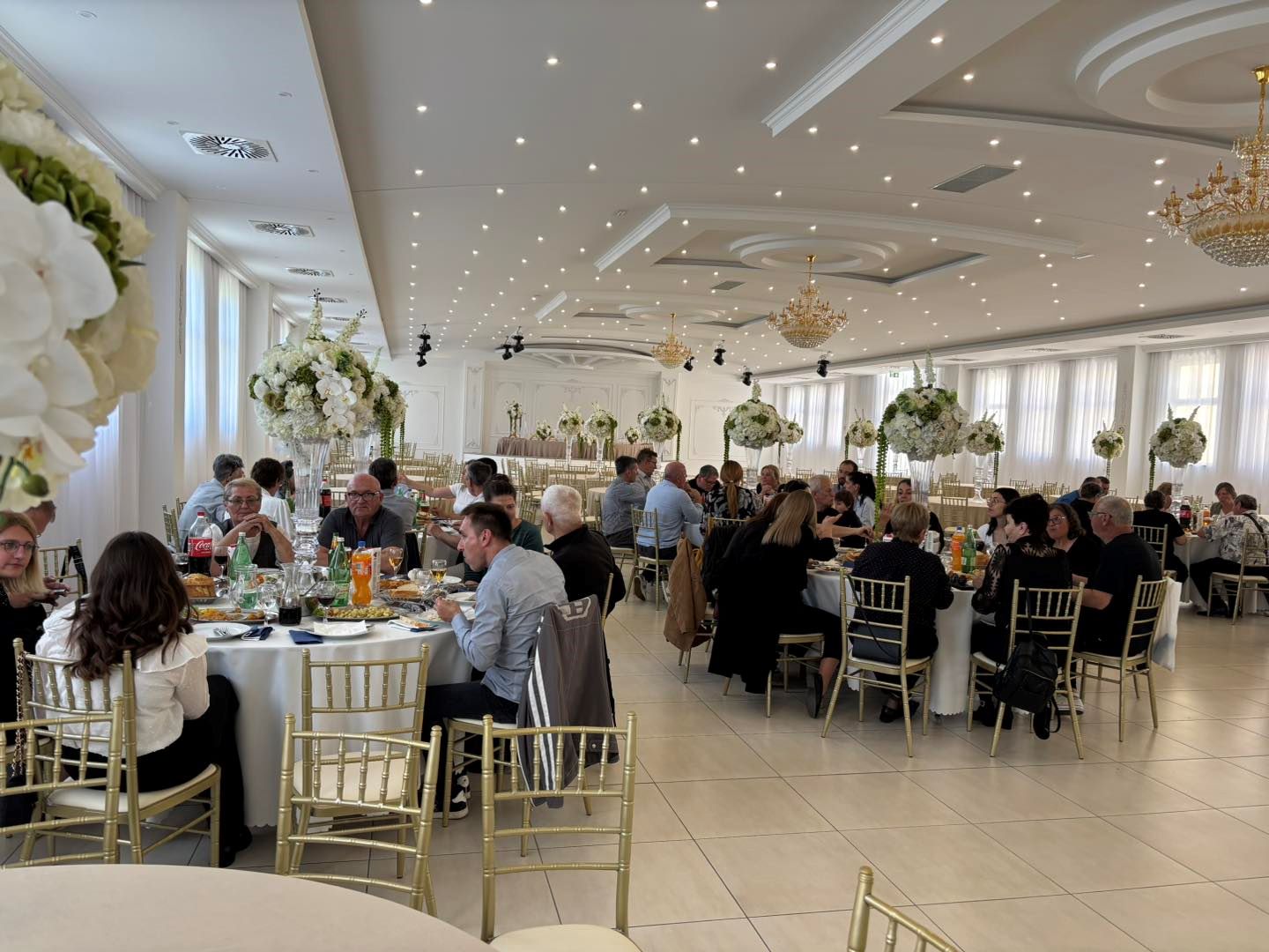 Na slici je prikazana svečana proslava u elegantnoj dvorani. Prostor je svijetao i prostran, s bijelim zidovima, visokim stropom ukrašenim rasvjetom i velikim lusterima koji dodatno naglašavaju svečani ugođaj. U dvorani se nalazi više okruglih stolova za kojima sjede uzvanici, većinom odrasli i stariji ljudi, odjeveni u polusvečanu i svečanu odjeću. Na stolovima su poslužena jela i pića, uključujući bezalkoholna pića, vino i vodu, što upućuje na organizirani ručak ili domjenak. Stolovi su dekorirani visokim cvjetnim aranžmanima u bijelim i zelenim tonovima, što prostoru daje profinjen i svečan izgled. Stolice su zlatne, a cijeli ambijent djeluje uredno i luksuzno. Ljudi za stolovima razgovaraju, jedu i druže se, što stvara dojam ugodne i opuštene atmosfere tijekom proslave ili obilježavanja nekog važnog događaja.