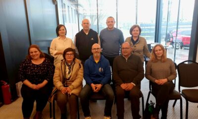 Na fotografiji je skupina od devet osoba okupljenih u zatvorenom prostoru, vjerojatno tijekom sastanka ili radnog susreta. Raspoređeni su u dva reda – pet osoba sjedi u prvom redu na stolicama, dok četiri osobe stoje iza njih. Prostor izgleda moderno i svijetlo, s velikim staklenim stijenkama kroz koje se vidi vanjski prostor, uključujući parkiralište i vozila. Rasvjeta je topla, a prostor djeluje uredno i poslovno. Osobe su odjevene u svakodnevnu, ali urednu odjeću – kombinaciju džempera, košulja i jakni. Većina njih gleda prema kameri s blagim osmijehom ili neutralnim izrazom lica, što odaje dojam formalnog, ali ugodnog okupljanja. U prvom redu, u sredini, nalazi se muškarac u plavoj majici s kapuljačom, dok su ostali oko njega raspoređeni simetrično. Iza njih stoje četiri osobe, također okrenute prema kameri. S lijeve strane uz zid vidi se i vatrogasni aparat. Cjelokupni dojam fotografije je profesionalan i kolegijalan, kao da prikazuje tim ili članove neke organizacije nakon sastanka ili događanja.