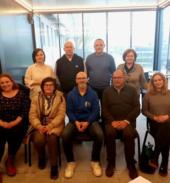 Na fotografiji je skupina od devet osoba okupljenih u zatvorenom prostoru, vjerojatno tijekom sastanka ili radnog susreta. Raspoređeni su u dva reda – pet osoba sjedi u prvom redu na stolicama, dok četiri osobe stoje iza njih. Prostor izgleda moderno i svijetlo, s velikim staklenim stijenkama kroz koje se vidi vanjski prostor, uključujući parkiralište i vozila. Rasvjeta je topla, a prostor djeluje uredno i poslovno. Osobe su odjevene u svakodnevnu, ali urednu odjeću – kombinaciju džempera, košulja i jakni. Većina njih gleda prema kameri s blagim osmijehom ili neutralnim izrazom lica, što odaje dojam formalnog, ali ugodnog okupljanja. U prvom redu, u sredini, nalazi se muškarac u plavoj majici s kapuljačom, dok su ostali oko njega raspoređeni simetrično. Iza njih stoje četiri osobe, također okrenute prema kameri. S lijeve strane uz zid vidi se i vatrogasni aparat. Cjelokupni dojam fotografije je profesionalan i kolegijalan, kao da prikazuje tim ili članove neke organizacije nakon sastanka ili događanja.