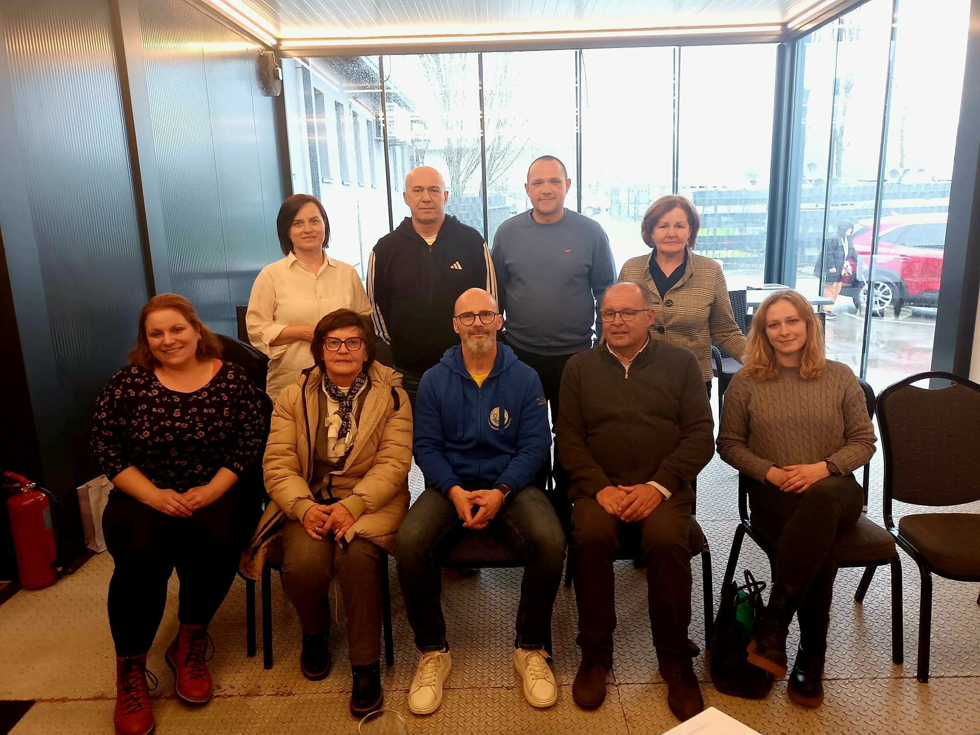 Na fotografiji je skupina od devet osoba okupljenih u zatvorenom prostoru, vjerojatno tijekom sastanka ili radnog susreta. Raspoređeni su u dva reda – pet osoba sjedi u prvom redu na stolicama, dok četiri osobe stoje iza njih. Prostor izgleda moderno i svijetlo, s velikim staklenim stijenkama kroz koje se vidi vanjski prostor, uključujući parkiralište i vozila. Rasvjeta je topla, a prostor djeluje uredno i poslovno. Osobe su odjevene u svakodnevnu, ali urednu odjeću – kombinaciju džempera, košulja i jakni. Većina njih gleda prema kameri s blagim osmijehom ili neutralnim izrazom lica, što odaje dojam formalnog, ali ugodnog okupljanja. U prvom redu, u sredini, nalazi se muškarac u plavoj majici s kapuljačom, dok su ostali oko njega raspoređeni simetrično. Iza njih stoje četiri osobe, također okrenute prema kameri. S lijeve strane uz zid vidi se i vatrogasni aparat. Cjelokupni dojam fotografije je profesionalan i kolegijalan, kao da prikazuje tim ili članove neke organizacije nakon sastanka ili događanja.