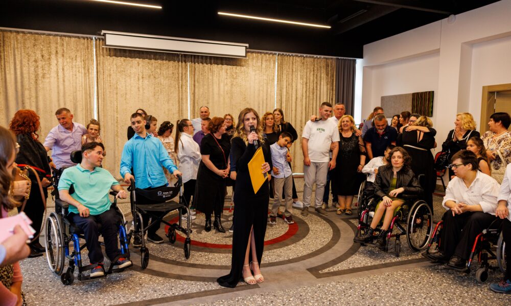 Na fotografiji je zabilježen trenutak s događaja u zatvorenom prostoru, najvjerojatnije tijekom modne revije ili sličnog društvenog okupljanja. U središtu pažnje nalazi se žena odjevena u elegantnu crnu haljinu koja drži mikrofon i žutu mapu, što upućuje na to da ima ulogu voditeljice ili organizatorice programa. Oko nje se nalazi veća skupina ljudi različite dobi, raspoređenih u polukrugu. Dio sudionika sjedi u invalidskim kolicima, dok ostali stoje, razgovaraju ili prate događanje. Atmosfera djeluje opušteno i emotivno, s vidljivim izrazima podrške, pažnje i međusobne povezanosti među prisutnima. Prostor je moderno uređen, s velikim zavjesama u pozadini i stropnom rasvjetom koja daje toplinu cijeloj sceni. Pod je ukrašen kružnim uzorkom koji dodatno naglašava središnju poziciju voditeljice. Fotografija odiše zajedništvom, inkluzijom i osjećajem zajedničkog slavlja