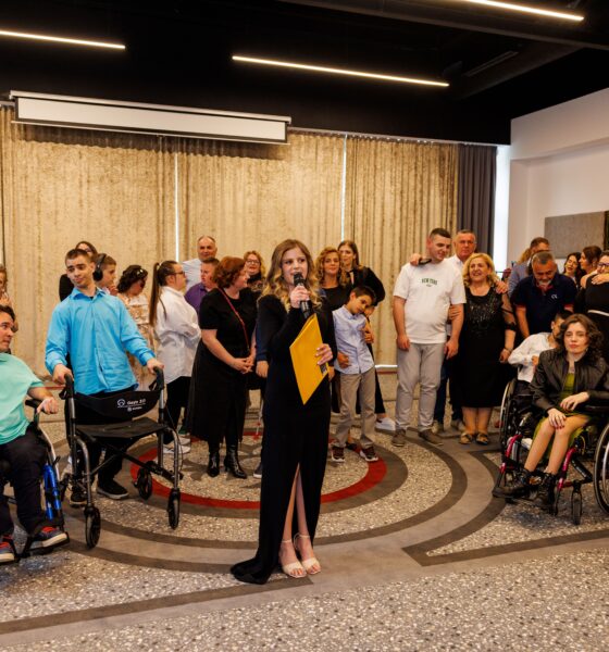 Na fotografiji je zabilježen trenutak s događaja u zatvorenom prostoru, najvjerojatnije tijekom modne revije ili sličnog društvenog okupljanja. U središtu pažnje nalazi se žena odjevena u elegantnu crnu haljinu koja drži mikrofon i žutu mapu, što upućuje na to da ima ulogu voditeljice ili organizatorice programa. Oko nje se nalazi veća skupina ljudi različite dobi, raspoređenih u polukrugu. Dio sudionika sjedi u invalidskim kolicima, dok ostali stoje, razgovaraju ili prate događanje. Atmosfera djeluje opušteno i emotivno, s vidljivim izrazima podrške, pažnje i međusobne povezanosti među prisutnima. Prostor je moderno uređen, s velikim zavjesama u pozadini i stropnom rasvjetom koja daje toplinu cijeloj sceni. Pod je ukrašen kružnim uzorkom koji dodatno naglašava središnju poziciju voditeljice. Fotografija odiše zajedništvom, inkluzijom i osjećajem zajedničkog slavlja