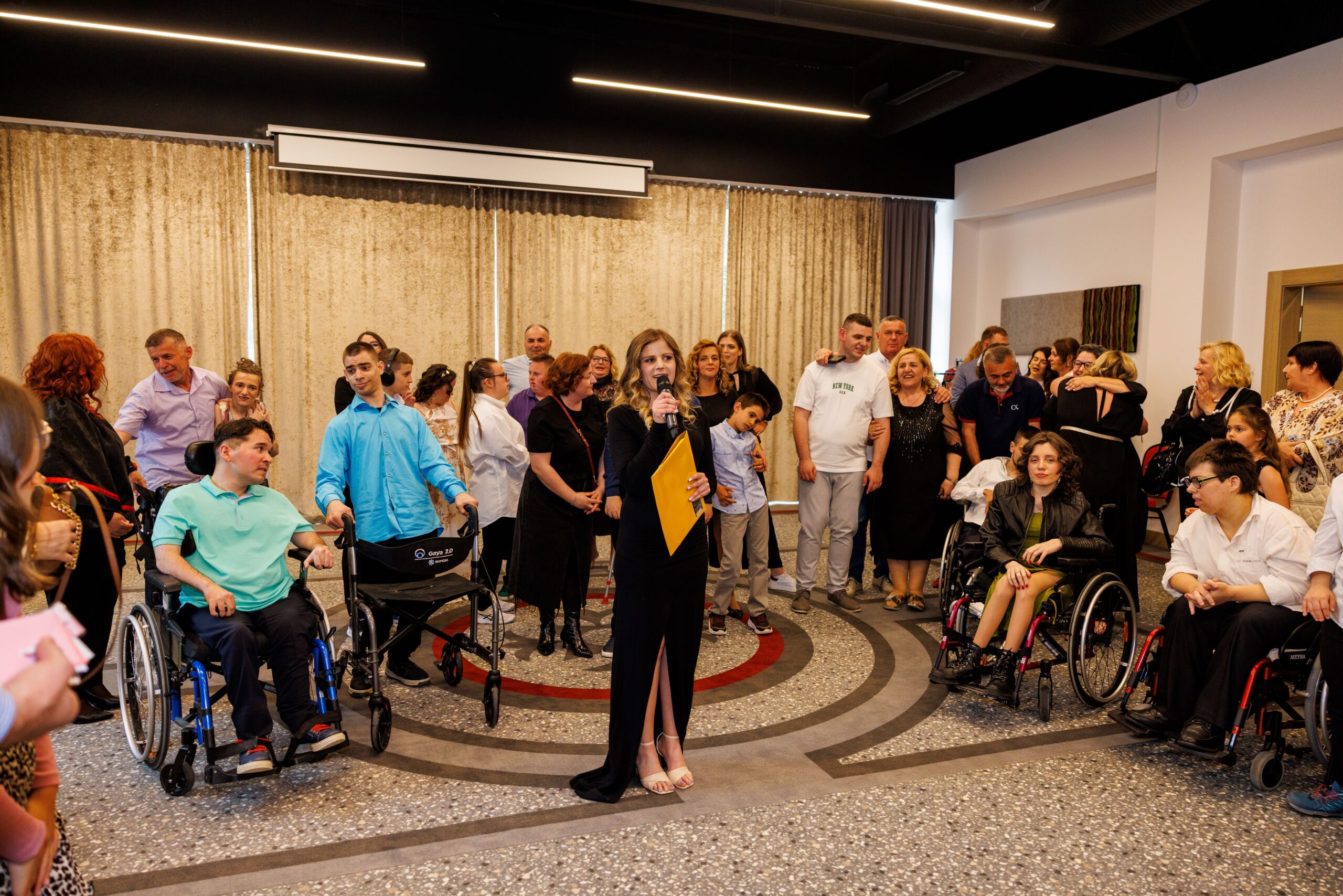 Na fotografiji je zabilježen trenutak s događaja u zatvorenom prostoru, najvjerojatnije tijekom modne revije ili sličnog društvenog okupljanja. U središtu pažnje nalazi se žena odjevena u elegantnu crnu haljinu koja drži mikrofon i žutu mapu, što upućuje na to da ima ulogu voditeljice ili organizatorice programa. Oko nje se nalazi veća skupina ljudi različite dobi, raspoređenih u polukrugu. Dio sudionika sjedi u invalidskim kolicima, dok ostali stoje, razgovaraju ili prate događanje. Atmosfera djeluje opušteno i emotivno, s vidljivim izrazima podrške, pažnje i međusobne povezanosti među prisutnima. Prostor je moderno uređen, s velikim zavjesama u pozadini i stropnom rasvjetom koja daje toplinu cijeloj sceni. Pod je ukrašen kružnim uzorkom koji dodatno naglašava središnju poziciju voditeljice. Fotografija odiše zajedništvom, inkluzijom i osjećajem zajedničkog slavlja