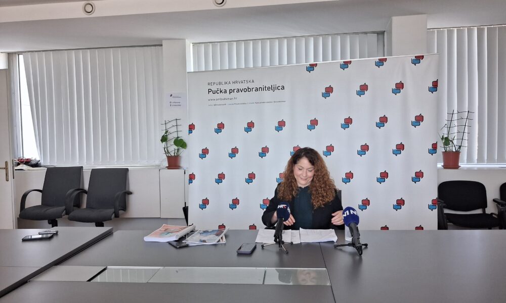 Fotografija prikazuje osobu kako sjedi za stolom, iza nje se nalazi neki baner.