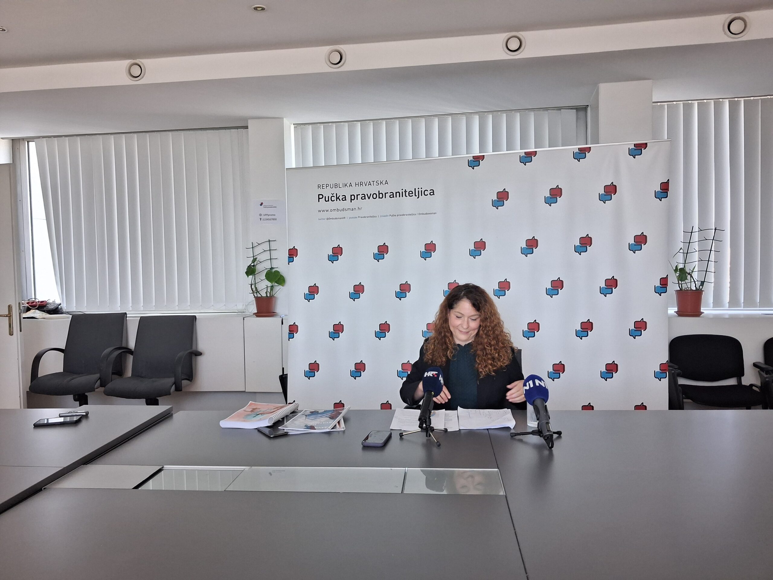 Fotografija prikazuje osobu kako sjedi za stolom, iza nje se nalazi neki baner.
