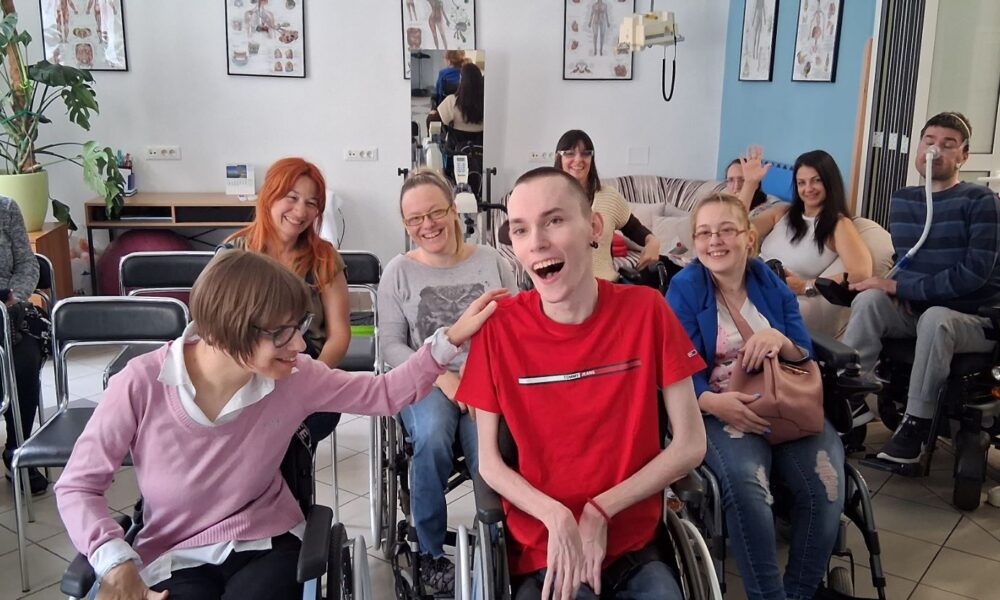 Na fotografiji je prikazana grupa ljudi u opuštenoj i veseloj atmosferi, okupljenih u prostoru koji nalikuje učionici ili terapijskoj/društvenoj sobi. U prvom planu nalaze se dvije osobe u invalidskim kolicima koje se smiju i međusobno komuniciraju – jedna osoba u ružičastom džemperu prijateljski dodiruje drugu osobu u crvenoj majici, koja se široko smije, što daje snažan dojam bliskosti i pozitivne energije. U pozadini se vidi još nekoliko sudionika, također nasmijanih i opuštenih, od kojih su neki u invalidskim kolicima. Jedna osoba maše prema kameri, dok drugi sjede i promatraju ili sudjeluju u razgovoru. S desne strane vidi se osoba koja stoji i pomaže muškarcu u kolicima koji koristi medicinsku opremu za disanje. Prostor je svijetao i uredan, s plakatima ljudske anatomije na zidovima i biljkom u kutu, što sugerira edukativno ili terapijsko okruženje. Cijela scena odiše toplinom, uključivošću i osjećajem zajedništva.
