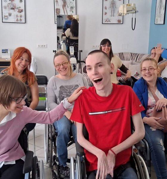 Na fotografiji je prikazana grupa ljudi u opuštenoj i veseloj atmosferi, okupljenih u prostoru koji nalikuje učionici ili terapijskoj/društvenoj sobi. U prvom planu nalaze se dvije osobe u invalidskim kolicima koje se smiju i međusobno komuniciraju – jedna osoba u ružičastom džemperu prijateljski dodiruje drugu osobu u crvenoj majici, koja se široko smije, što daje snažan dojam bliskosti i pozitivne energije. U pozadini se vidi još nekoliko sudionika, također nasmijanih i opuštenih, od kojih su neki u invalidskim kolicima. Jedna osoba maše prema kameri, dok drugi sjede i promatraju ili sudjeluju u razgovoru. S desne strane vidi se osoba koja stoji i pomaže muškarcu u kolicima koji koristi medicinsku opremu za disanje. Prostor je svijetao i uredan, s plakatima ljudske anatomije na zidovima i biljkom u kutu, što sugerira edukativno ili terapijsko okruženje. Cijela scena odiše toplinom, uključivošću i osjećajem zajedništva.