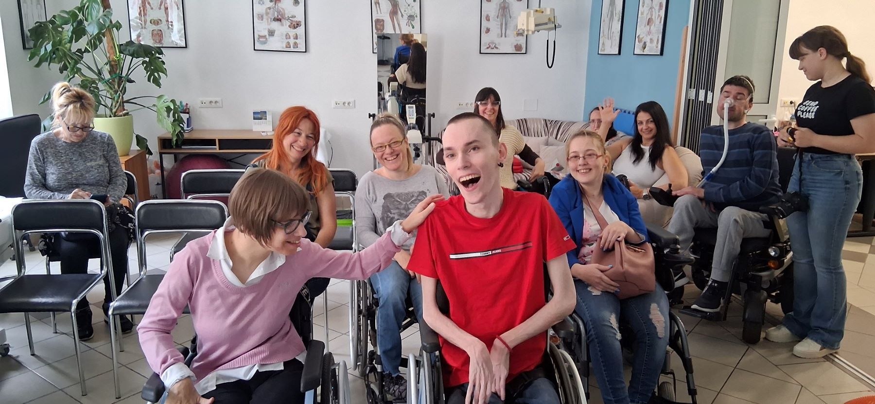 Na fotografiji je prikazana grupa ljudi u opuštenoj i veseloj atmosferi, okupljenih u prostoru koji nalikuje učionici ili terapijskoj/društvenoj sobi. U prvom planu nalaze se dvije osobe u invalidskim kolicima koje se smiju i međusobno komuniciraju – jedna osoba u ružičastom džemperu prijateljski dodiruje drugu osobu u crvenoj majici, koja se široko smije, što daje snažan dojam bliskosti i pozitivne energije. U pozadini se vidi još nekoliko sudionika, također nasmijanih i opuštenih, od kojih su neki u invalidskim kolicima. Jedna osoba maše prema kameri, dok drugi sjede i promatraju ili sudjeluju u razgovoru. S desne strane vidi se osoba koja stoji i pomaže muškarcu u kolicima koji koristi medicinsku opremu za disanje. Prostor je svijetao i uredan, s plakatima ljudske anatomije na zidovima i biljkom u kutu, što sugerira edukativno ili terapijsko okruženje. Cijela scena odiše toplinom, uključivošću i osjećajem zajedništva.