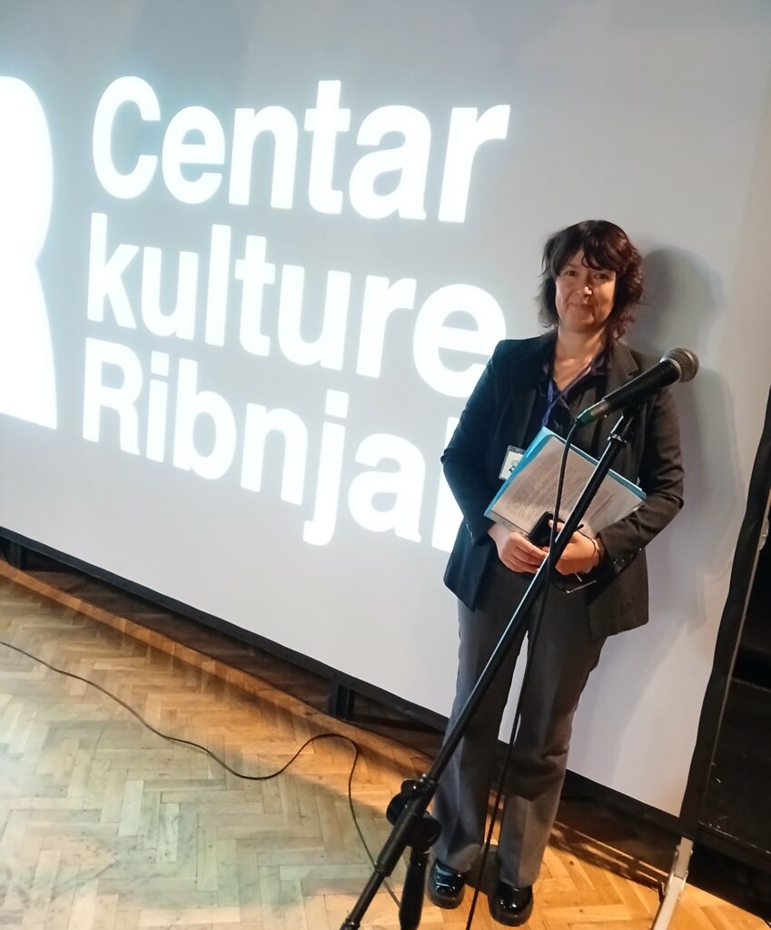 Žena stoji uz mikrofon ispred projekcije s natpisom 'Centar kulture Ribnjak', drži dokumente.