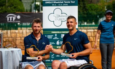 Fotograafija prikazuje dvoje ljudi u kolicima koji drže nagrade u jednoj, a pozdravljaju se s drugom rukom. Oni se nalaze na zemljanoj površini nekog teniskog terena.