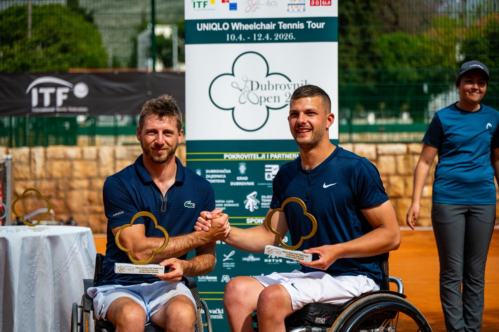 Fotograafija prikazuje dvoje ljudi u kolicima koji drže nagrade u jednoj, a pozdravljaju se s drugom rukom. Oni se nalaze na zemljanoj površini nekog teniskog terena.