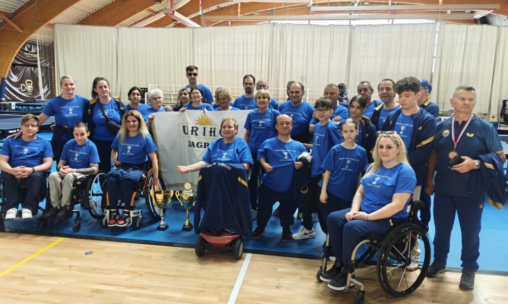 Fotografija prikazuje skupinu ljudi koja je osvojila pehar na nekom prvenstvu. Neki od njih su u kolicima, dok drugi stoje na nogama. Između njih je i zastava na kojoj piše 'URIHO Zagreb'.