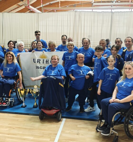 Fotografija prikazuje skupinu ljudi koja je osvojila pehar na nekom prvenstvu. Neki od njih su u kolicima, dok drugi stoje na nogama. Između njih je i zastava na kojoj piše 'URIHO Zagreb'.