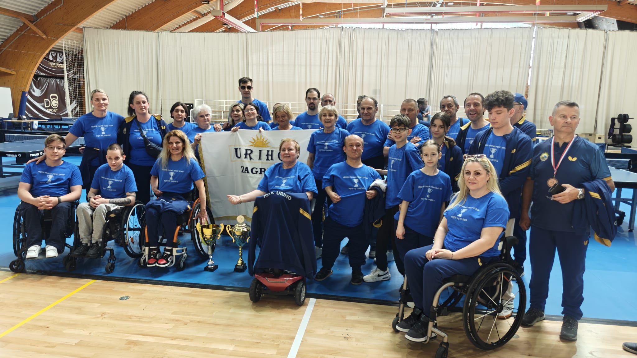 Fotografija prikazuje skupinu ljudi koja je osvojila pehar na nekom prvenstvu. Neki od njih su u kolicima, dok drugi stoje na nogama. Između njih je i zastava na kojoj piše 'URIHO Zagreb'.