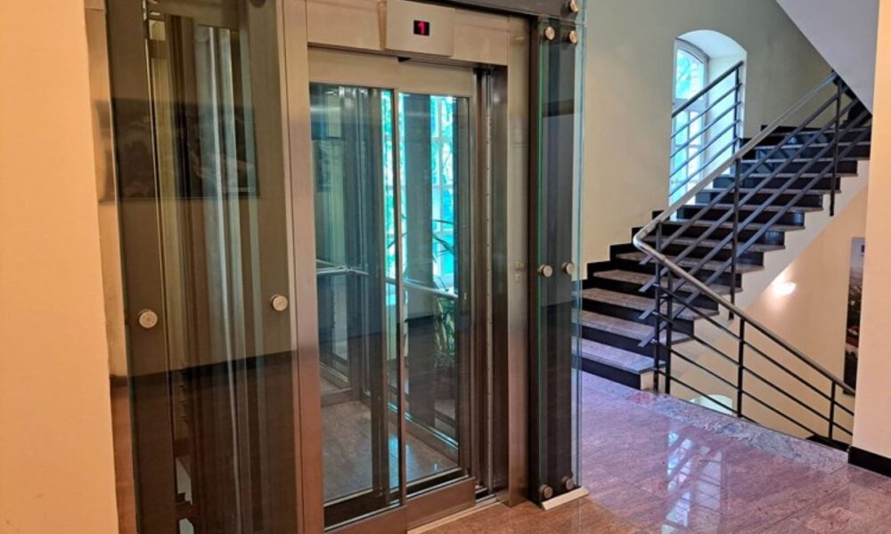 Na fotografiji se vidi moderno stakleno dizalo smješteno unutar stubišta zgrade. Dizalo ima prozirne stijenke i metalnu konstrukciju, što omogućuje pogled na unutrašnjost i okolni prostor. Uz dizalo se nalaze stepenice s tamnom metalnom ogradom koje vode na više katove. Pod je obložen sjajnim kamenom, a prostor djeluje uredno, svijetlo i suvremeno uređen.